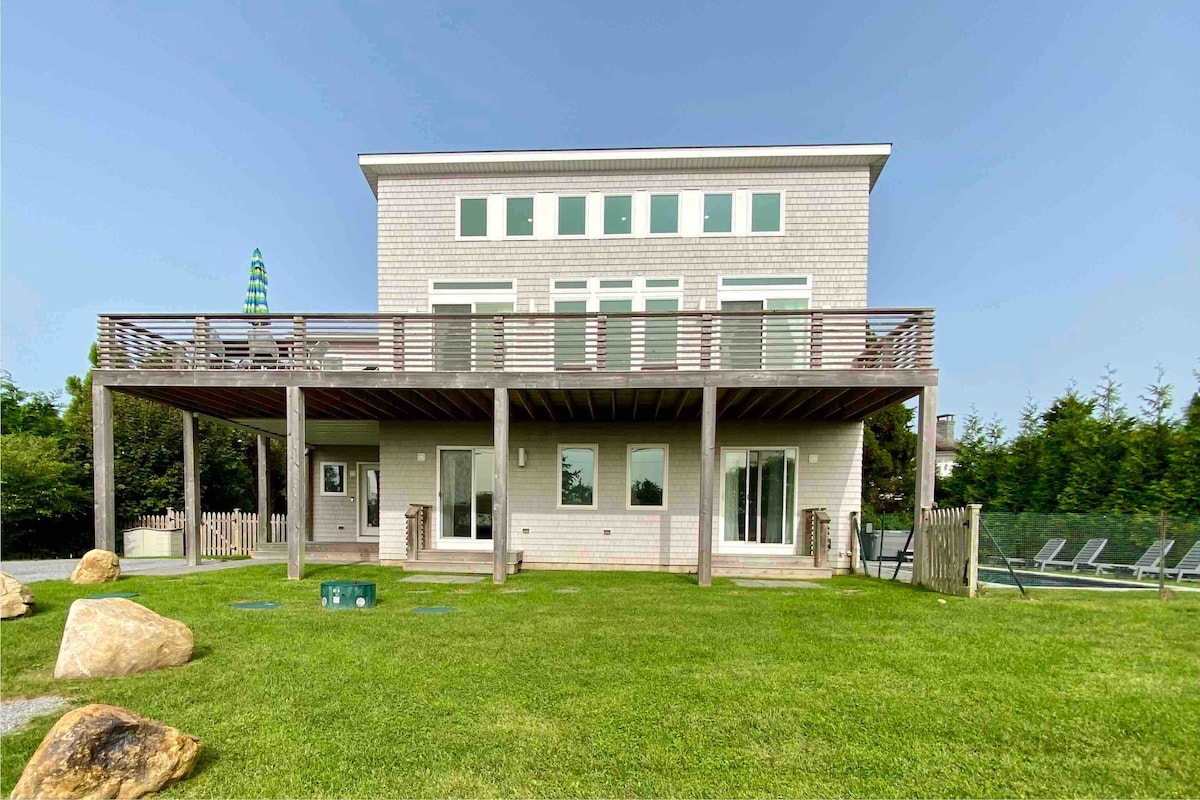 The beach house features a spacious second-level deck with ample seating and views of the surroundings. Large glass doors provide access to the yard, which is bordered by a grassy area and includes a hot tub. The exterior showcases modern architecture with light-colored siding.