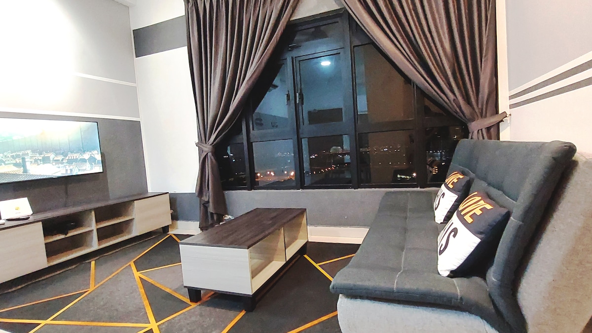 A modern living area is showcased, featuring a dark gray sofa adorned with decorative pillows. A sleek coffee table sits in front of the sofa, while a large window offers views of the city skyline at night, framed by dark curtains.