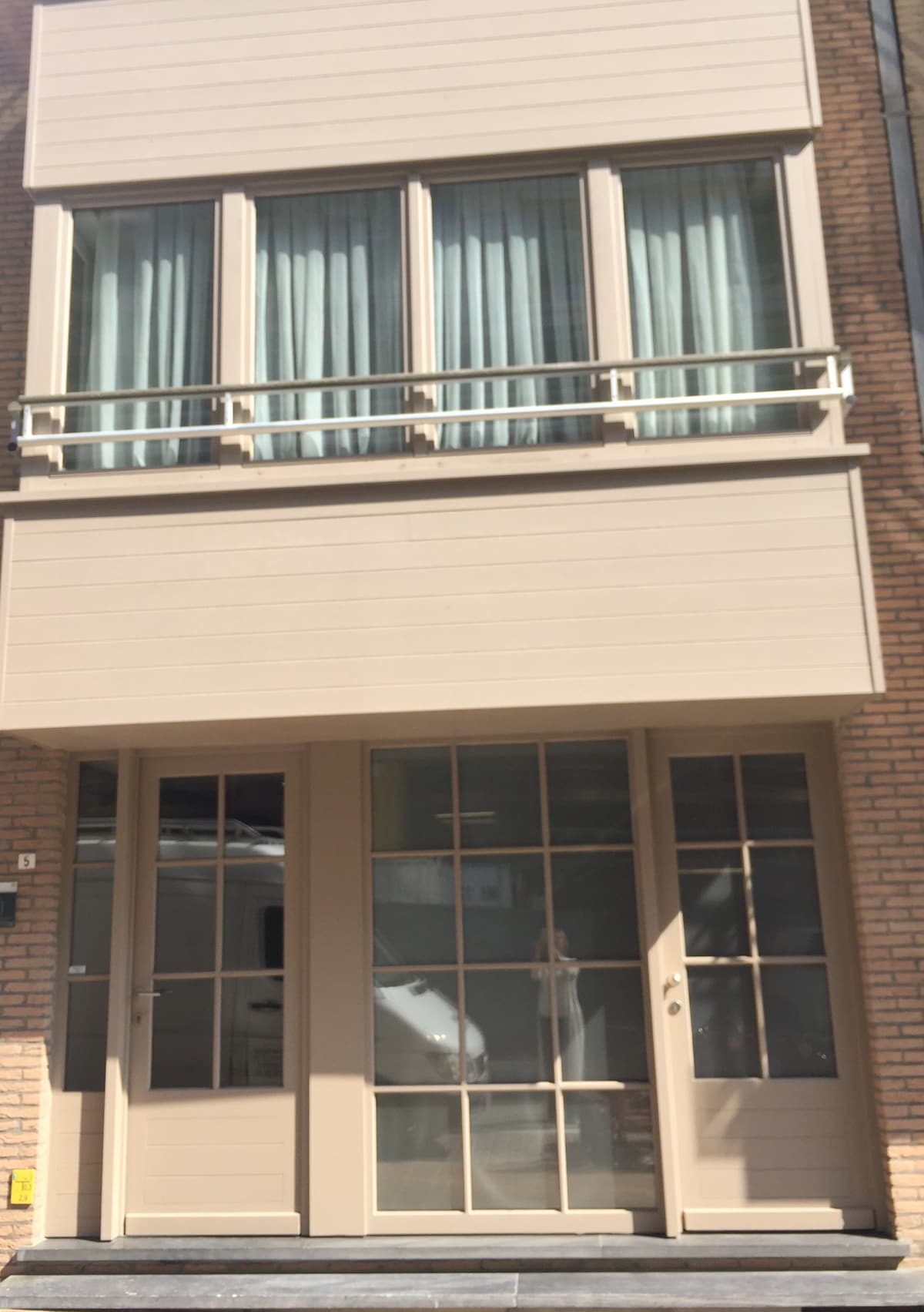 The exterior of the building features a warm beige facade with large glass doors framed by two sets of windows adorned with pale blue curtains. A small balcony overhangs the entrance, providing a welcoming touch to the inviting structure.