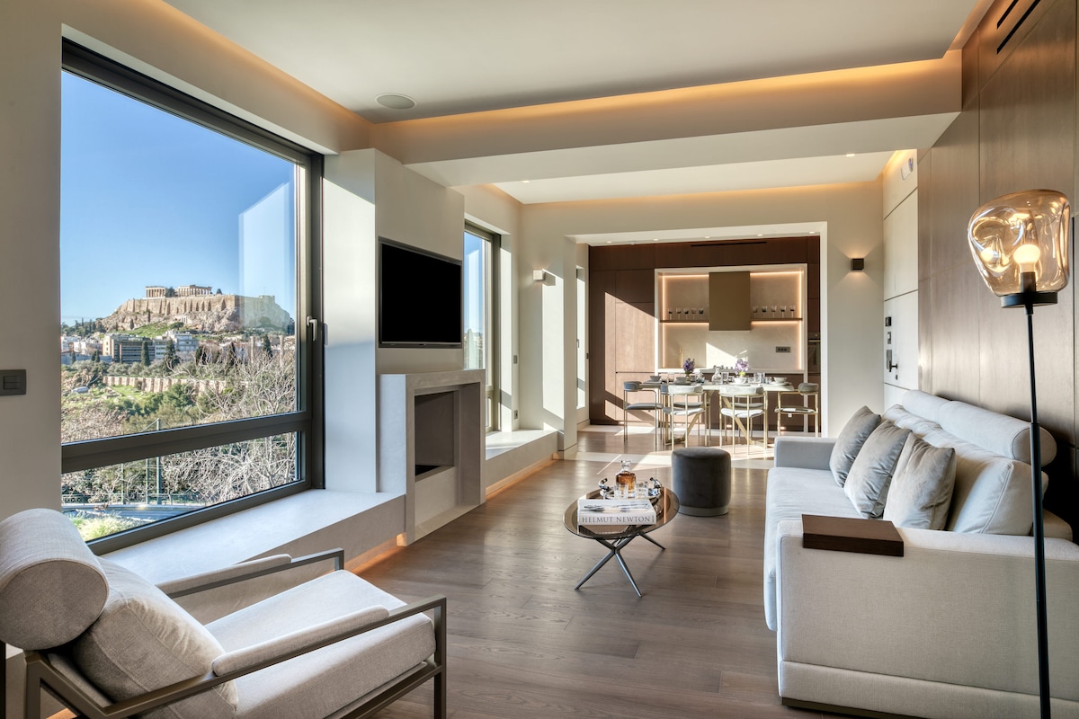 A spacious living area features large floor-to-ceiling windows, offering a view of the Acropolis. Modern furnishings include a comfortable sofa and an armchair, along with a coffee table. Natural light fills the room, highlighting the wooden floors and elegant interior design.