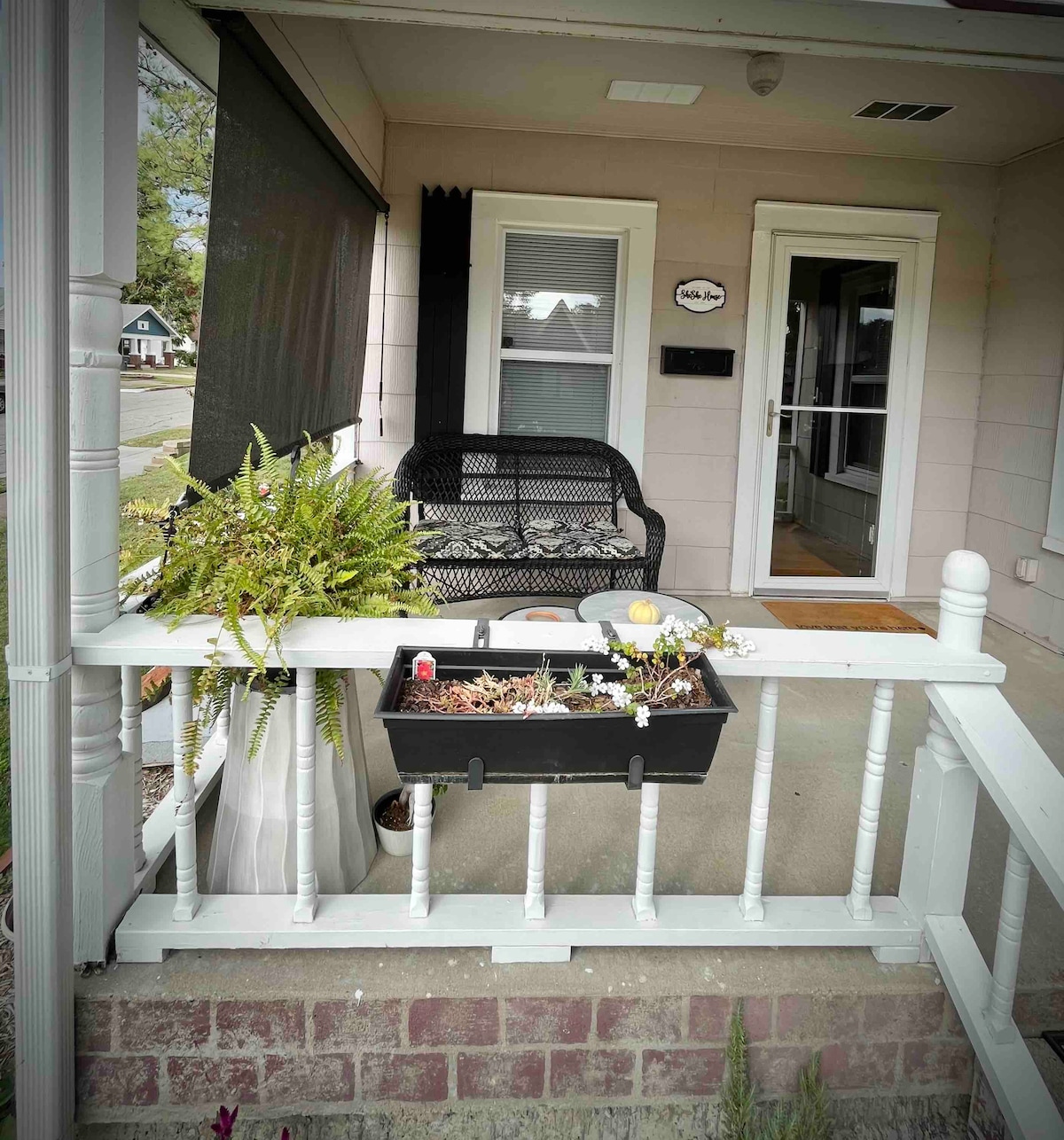 A covered front porch features a black wicker bench, providing a comfortable seating area. Potted plants are arranged on the railing, adding greenery. A welcome sign is displayed beside the door, enhancing the inviting ambiance of the entryway.
