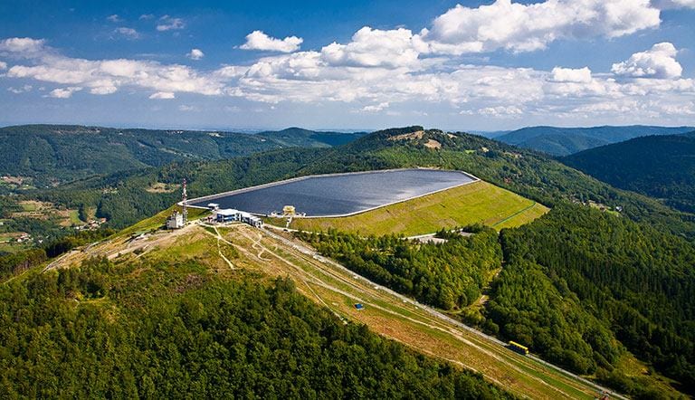 A sweeping view captures a solar panel installation on a hillside, surrounded by lush green forests and distant rolling mountains. The scene showcases the harmony between nature and renewable energy, with a clear blue sky adorned with scattered clouds enhancing the landscape.