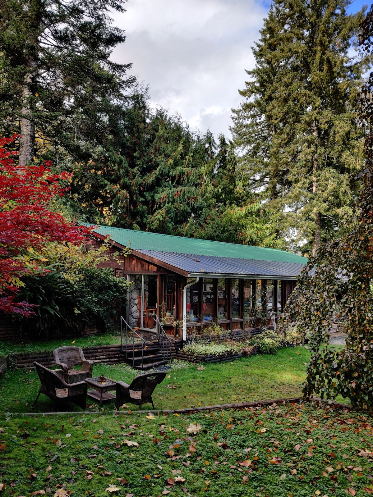 A charming exterior view of the property is framed by lush greenery and vibrant autumn leaves. The structure features large glass windows, allowing for natural light, while a set of four patio chairs surrounds a small table on the grassy area in front.
