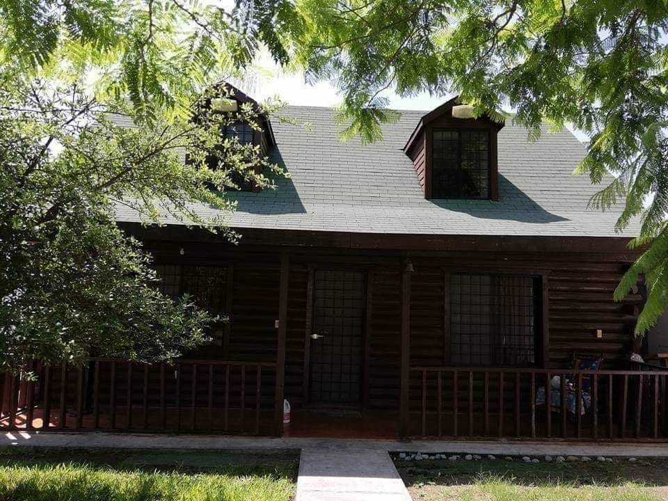 A rustic cabin is framed by lush greenery, featuring a distinctive wooden exterior. Two dormer windows enhance the charm of the sloped roof. A welcoming pathway leads to the entrance, flanked by a wooden railing, creating an inviting atmosphere.