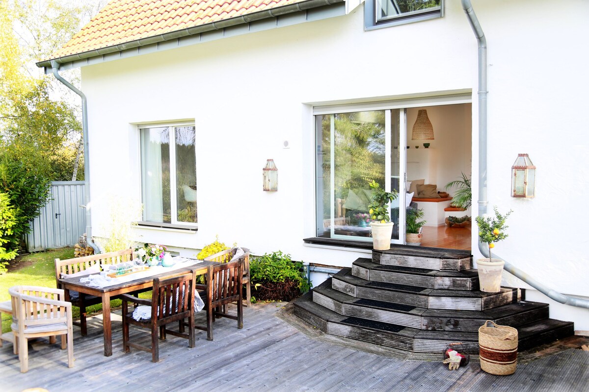 An outdoor terrace features a large wooden dining table with several chairs, surrounded by greenery. Steps lead up to the house, where large windows provide a view into the living area. Potted plants are placed decoratively on both the terrace and at the entrance.