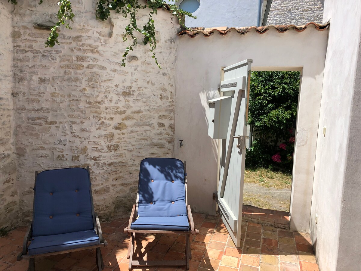 An outdoor area features two blue lounge chairs positioned on terracotta tiles. Sunlight casts gentle shadows, while a stone wall provides texture. An open door leads to a garden area with greenery, enhancing the tranquil setting.