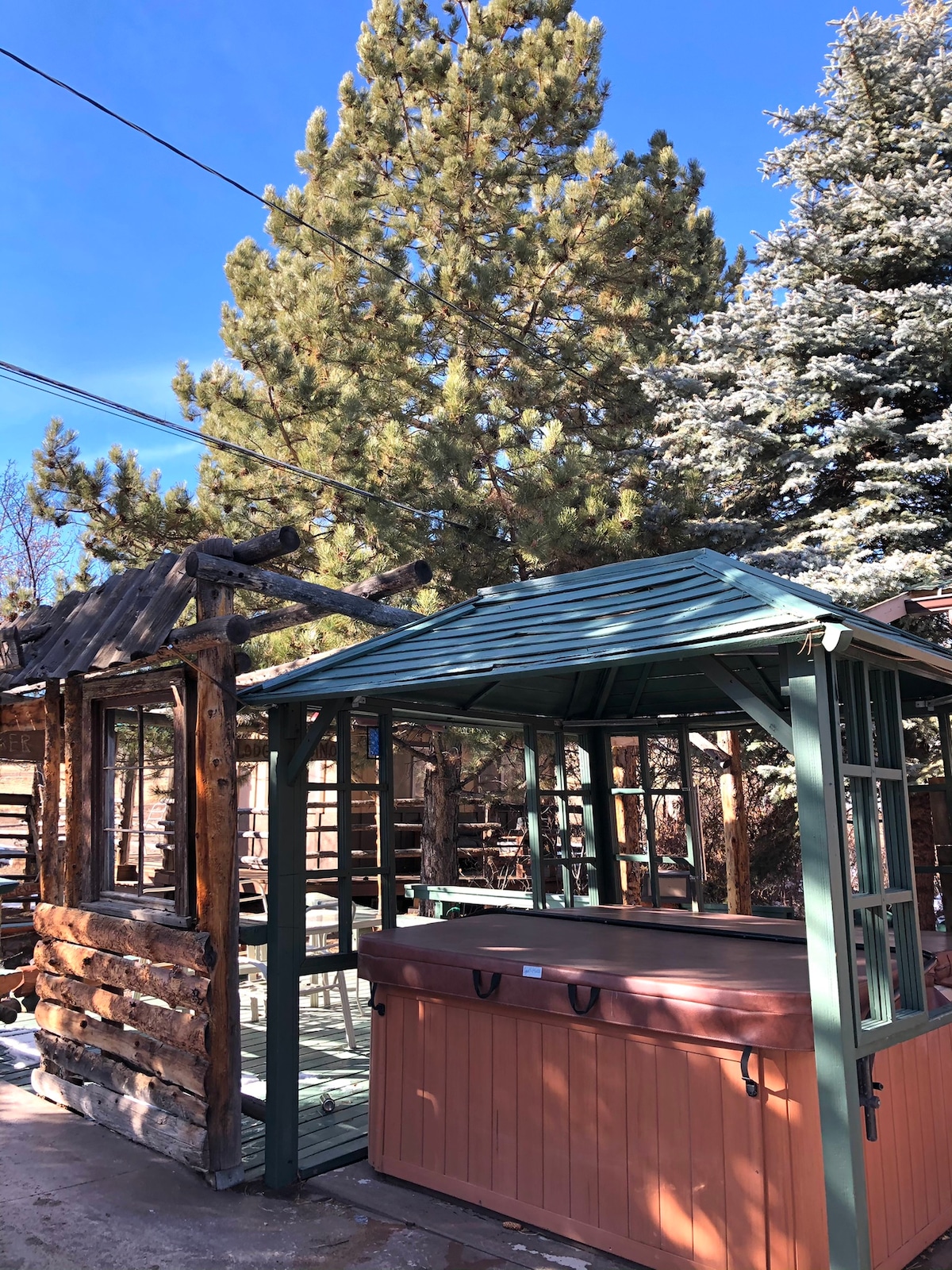 A rustic outdoor hot tub is framed by a wooden gazebo, providing a welcoming space for relaxation. Towering pine trees create a natural backdrop, while clear blue skies enhance the outdoor setting.
