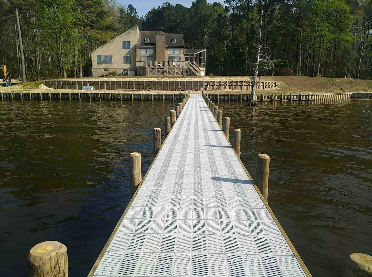 A long, white dock extends over the water, leading to a spacious vacation home set against a backdrop of lush greenery. The house features multiple levels and large windows, offering views of the river from various angles. The area is calm and serene.