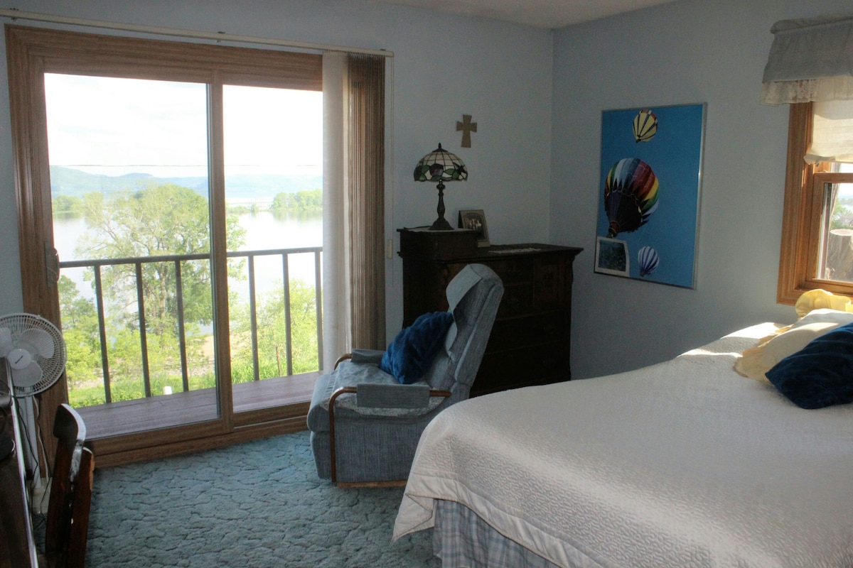 A serene bedroom features a spacious bed with light bedding, complemented by a blue carpet. A rocking chair is positioned near a sliding glass door, providing views of the river and trees outside. An art piece with colorful balloons adds a touch of decor.