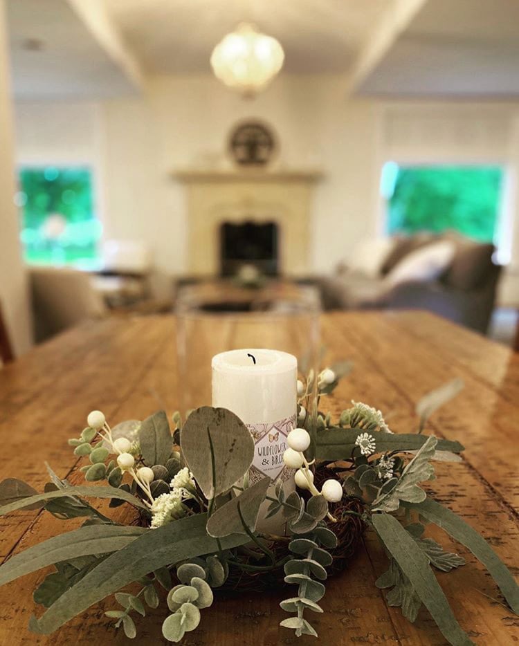 A wooden dining table is set with a decorative centerpiece featuring a white candle surrounded by greenery and small white flowers. In the background, a cozy living area is revealed, including a fireplace and comfortable seating, with ample natural light coming through large windows.