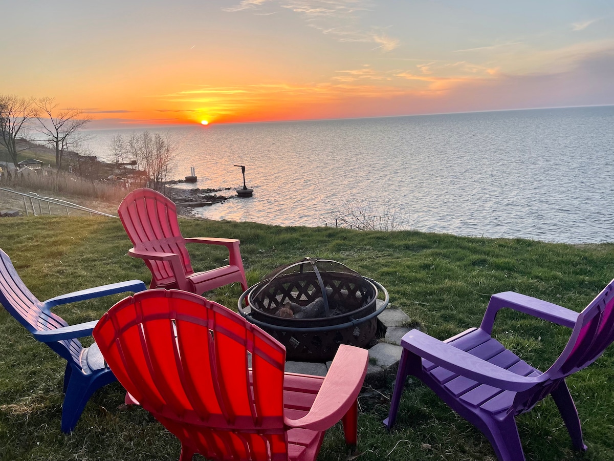 Colorful Adirondack chairs surround a fire pit, positioned on a grassy area near the water's edge. The sun is setting over the lake, casting warm hues across the sky and reflecting on the water. A serene atmosphere is created by the tranquil lake views.