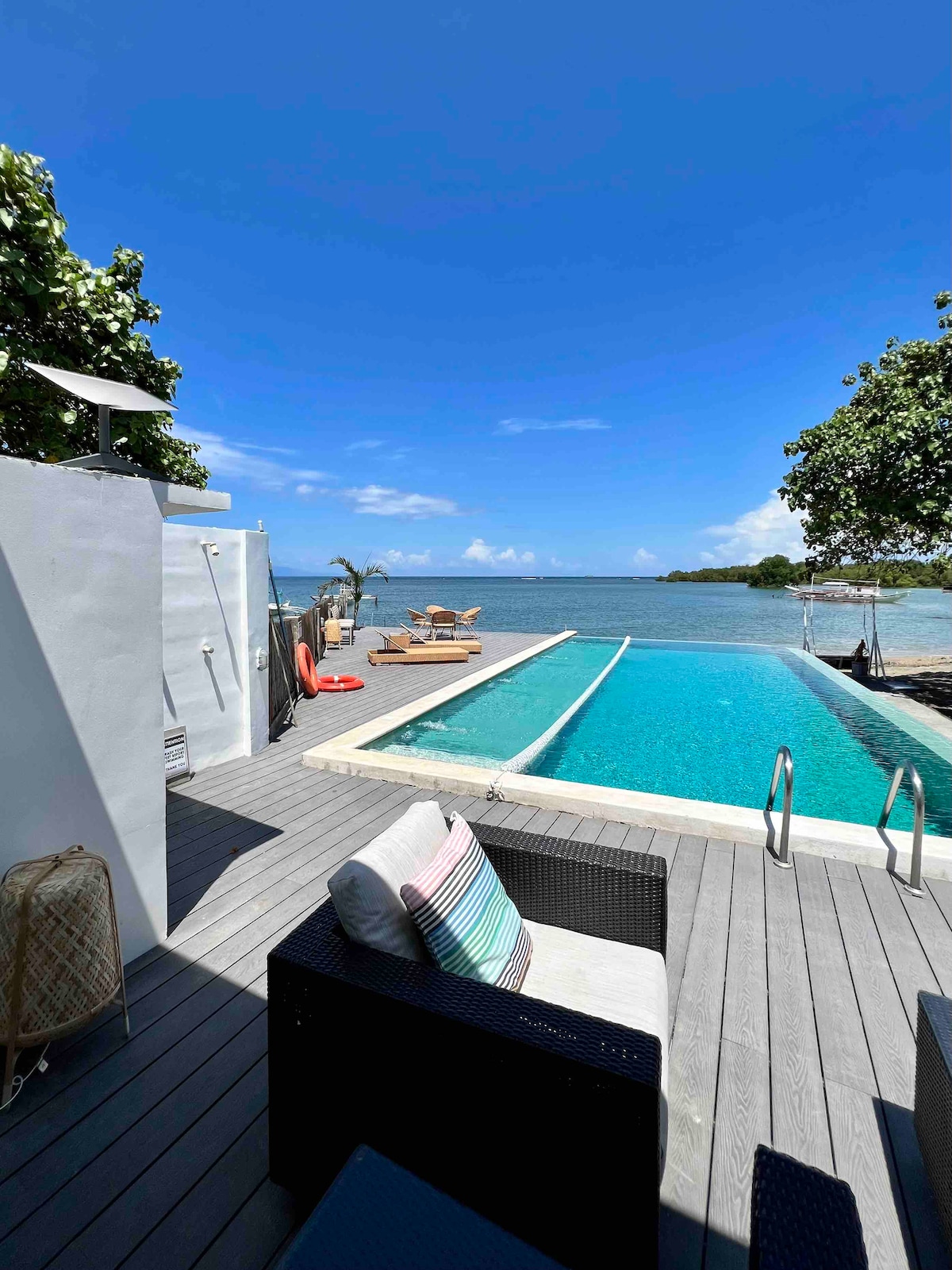 An expansive infinity pool is seen merging with the sea, framed by a wooden deck. Comfortable lounge chairs sit near the pool, surrounded by lush greenery and offering views of the water under a bright blue sky.