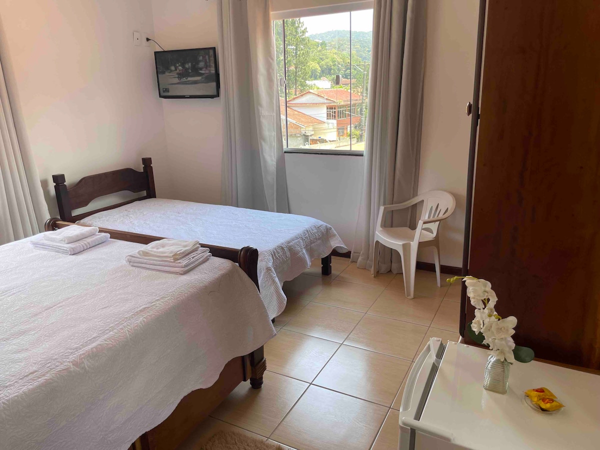 The room features two wooden beds with white linens, arranged beside a window with drapes that allows natural light. A small television is mounted on the wall, and a plastic chair is positioned nearby. A dresser and a vase with flowers are visible as well.