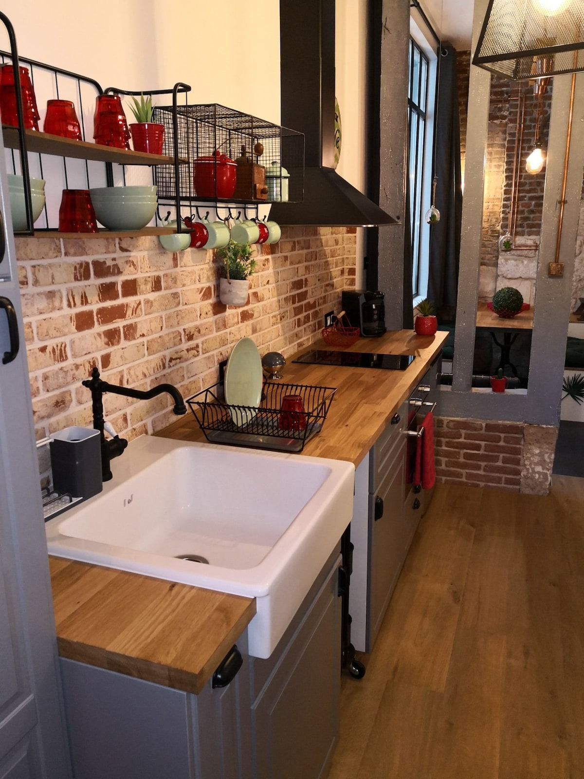 A modern kitchen features a spacious wooden countertop complemented by a farmhouse sink. Open shelving displays colorful dishware against a backdrop of exposed brick walls. Contemporary appliances and decorative accents can be seen, creating a functional and inviting cooking space.