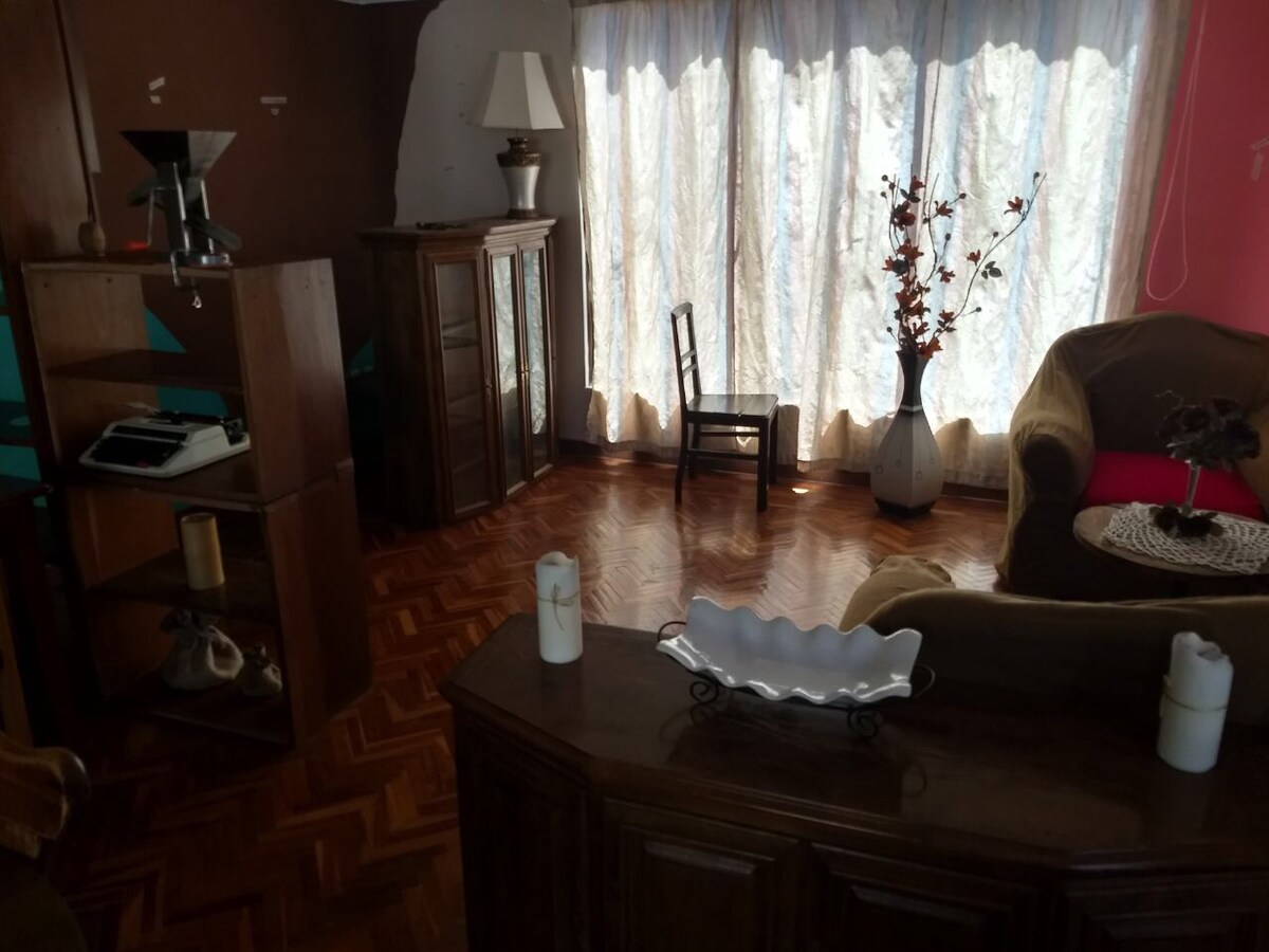 A comfortable living space features a mix of furniture, including a wooden cabinet, a small chair, and a decorative vase with faux flowers. Natural light filters through sheer curtains, highlighting the warm tones of the polished wood flooring.