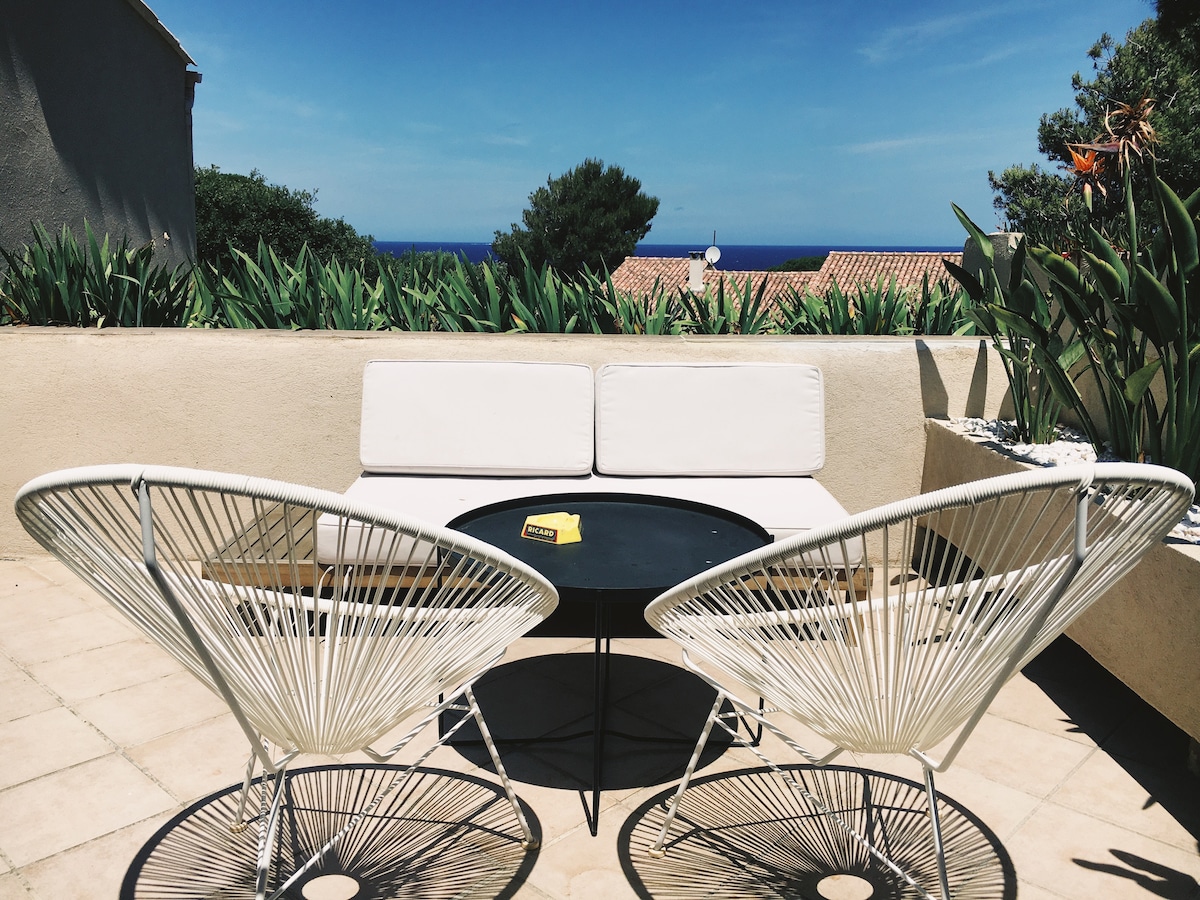 An outdoor seating area is featured, furnished with two white lounge chairs and a round black table. Lush greenery surrounds the space, and the distant view of blue ocean water complements the relaxing atmosphere.