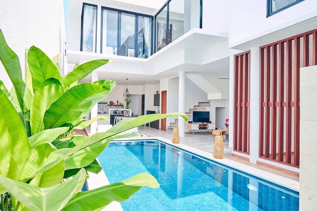 A private pool is surrounded by tropical greenery, with a smooth stone deck. Large glass doors offer a view of the airy living area, which includes a modern TV setup and furniture. The villa's design emphasizes open space, natural light, and minimalist architecture.