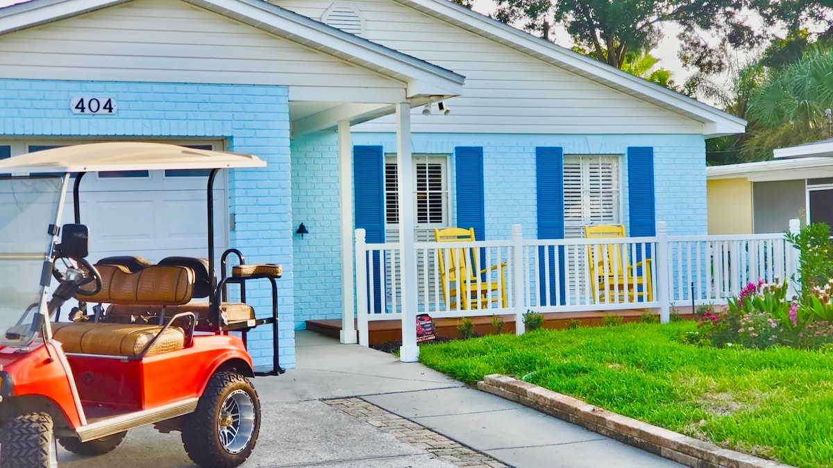 The exterior of the bungalow features a charming blue brick facade, complemented by white trim and shutters. A red electric buggy is parked in the foreground, while cozy yellow chairs are positioned on the porch behind a white railing, surrounded by well-maintained greenery.