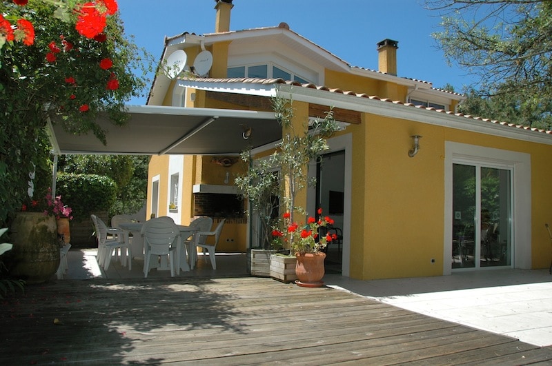 A sunny outdoor terrace is visible, featuring a large table with several white chairs. Flower pots and climbing plants add greenery to the surroundings. The exterior walls of the house are painted yellow, with glass doors providing access to the interior.