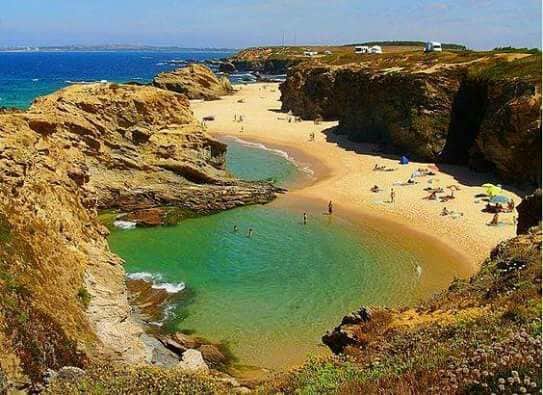 A coastal view captures a sandy beach bordered by rugged rocks. Gentle waves lap at the shoreline, and visitors can be seen enjoying the water, some under colorful umbrellas. The vibrant green water contrasts with the golden sand, creating a serene natural environment.