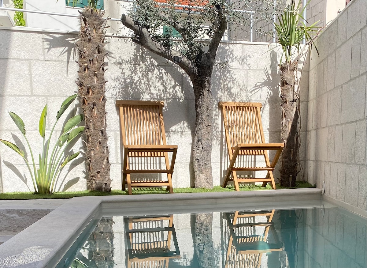 Two wooden chairs are positioned beside a tranquil pool, with palm trees and lush greenery enhancing the outdoor ambiance. The stone wall surrounding the area provides privacy and a natural feel, while reflections in the water create a serene atmosphere.