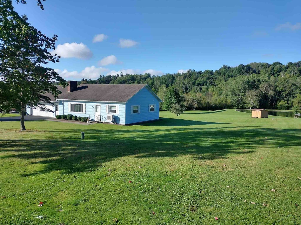 A renovated ranch home is presented in a serene country setting, surrounded by ample grassy areas. Trees frame the property, which features views of woods and a pond in the background. The house's blue exterior complements the clear sky and natural landscape.