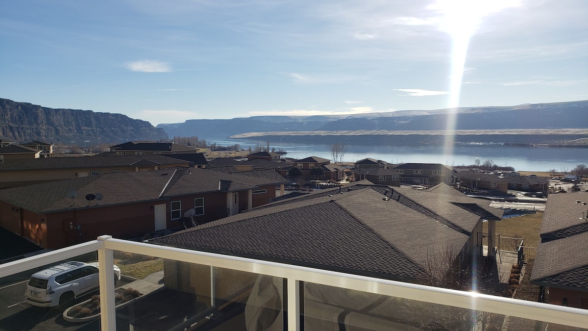 A wide view from the balcony captures a serene landscape featuring rolling hills and a calm water body reflecting the sky. The image includes nearby rooftops, adding depth to the scene, which is illuminated by soft sunlight.