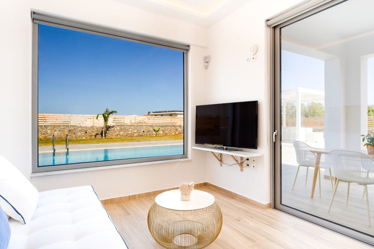 A bright living area is showcased with a large window offering a view of the private pool and garden. A modern white sofa is positioned beside a round table, and a flat-screen TV is mounted on the wall, providing entertainment options. Natural light fills the space.