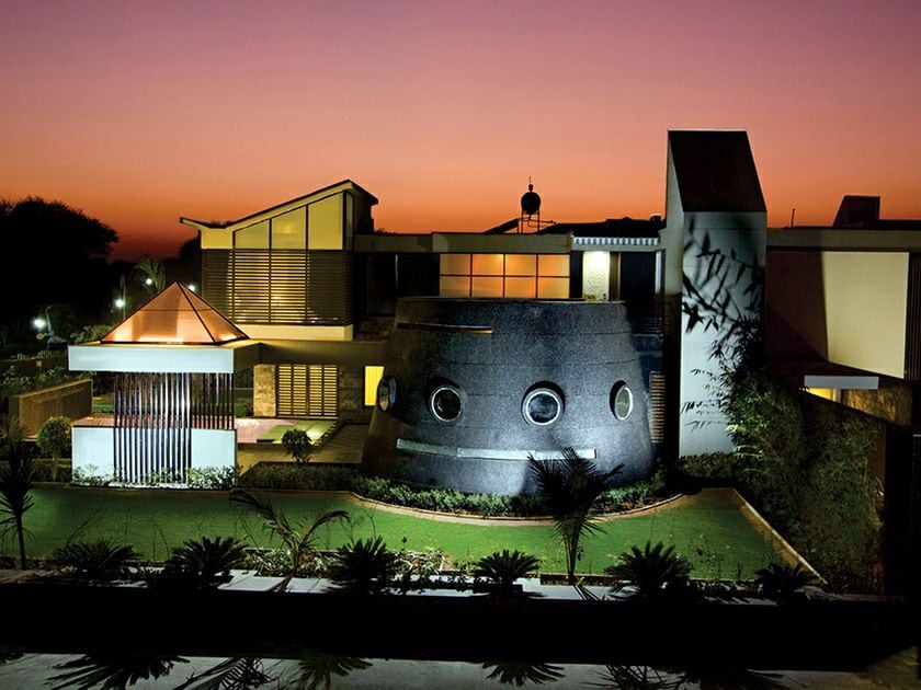 The villa is presented with a unique architectural design, featuring circular windows and a striking silhouette against a sunset sky. Landscaped greenery surrounds the structure, accentuating its contemporary feel. Soft lighting highlights the home’s features, inviting a sense of tranquility in the evening atmosphere.