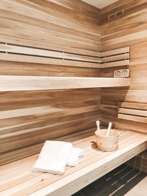 A wooden sauna interior is displayed, featuring slatted wooden walls and benches. A small wooden bucket and ladle are positioned on the bench, alongside a folded white towel. Controls for temperature are clearly visible on the wall.