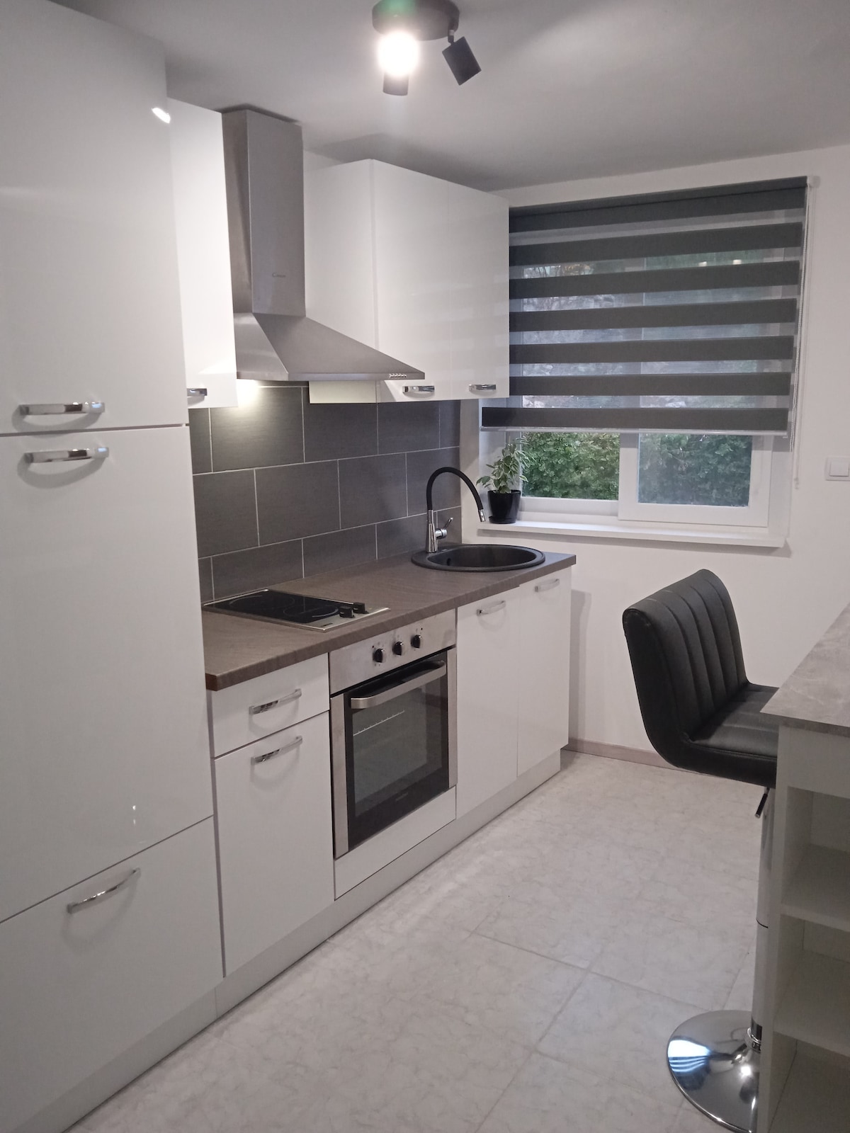 A modern kitchen features sleek white cabinetry and a gray-tiled backsplash. Equipped with a stove, oven, and sink, the space also includes a bar counter with stools. Natural light filters through the window adorned with striped blinds, illuminating the functional design.