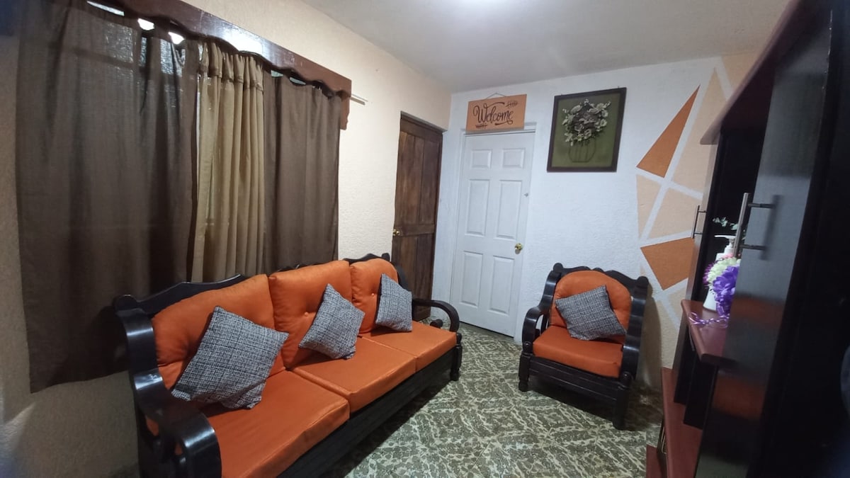 A living area features a dark wooden sofa set adorned with orange cushions and gray throw pillows, complemented by a matching armchair. Natural light filters through brown drapes, illuminating the textured floor, while a decorative wall piece adds character to the room.