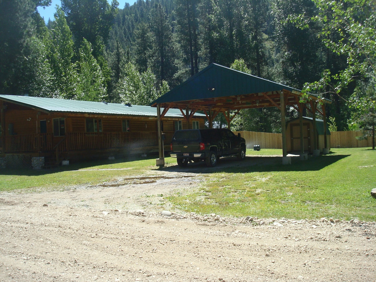 A charming log cabin with a green metal roof is surrounded by trees, creating a serene environment. A spacious covered carport accommodates a vehicle, while a well-manicured lawn offers an inviting space for outdoor activities.