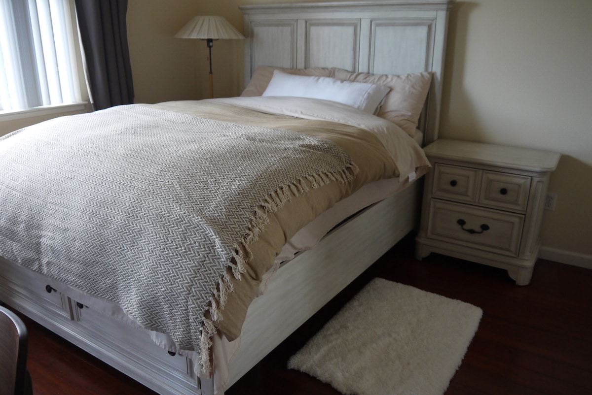 A spacious bed with a soft, neutral-toned blanket is positioned against a light-colored headboard. A nightstand with drawers is located beside the bed, and a plush area rug is placed on the polished wooden floor. Natural light filters through the window, enhancing the cozy ambiance.