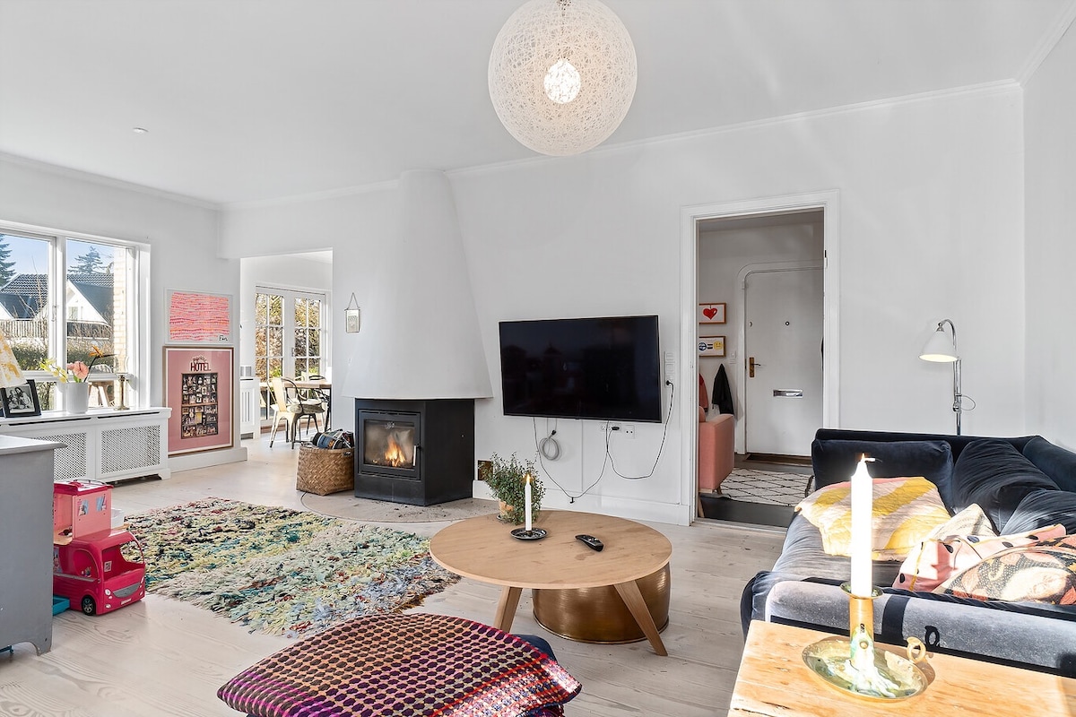 A bright living room features a cozy seating area with a dark sofa and a round coffee table. A patterned rug adds warmth to the light wood flooring. A modern chandelier hangs above, while large windows allow natural light to fill the space. A fireplace is visible, enhancing the inviting atmosphere.