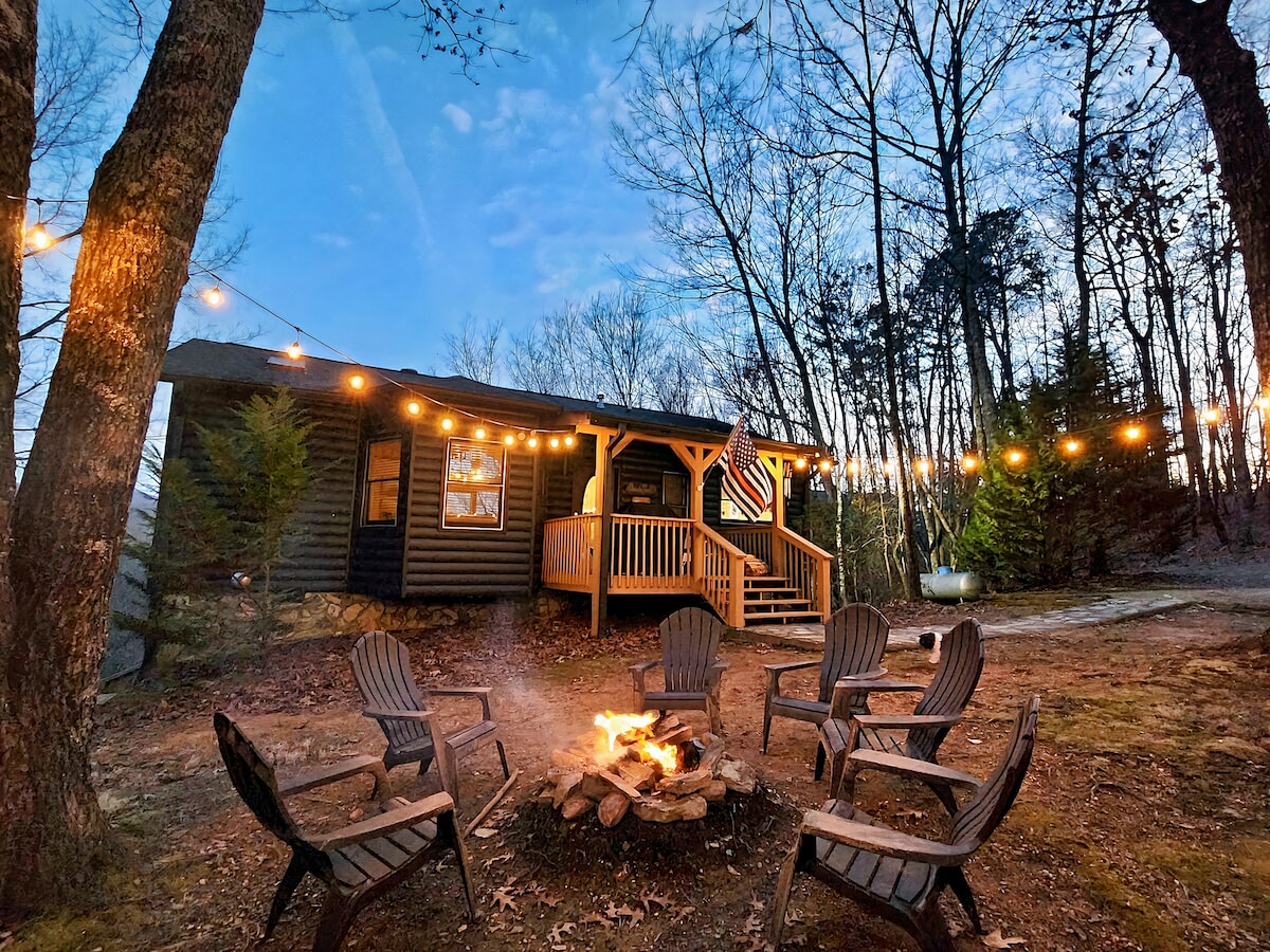 An outdoor fire pit is surrounded by several Adirondack chairs, creating a cozy space for gatherings. Soft string lights are draped overhead, illuminating the area against a twilight backdrop. The cabin features a welcoming porch visible in the background, surrounded by trees.