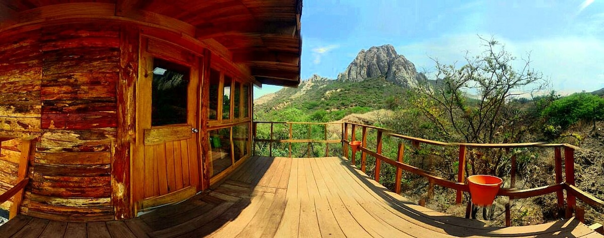 A wooden deck surrounds the cabin, offering expansive views of the surrounding natural landscape and distant rock formations. The area is bathed in natural light, highlighting the wood grain of the structure. A sense of openness is created through the large windows and railing.