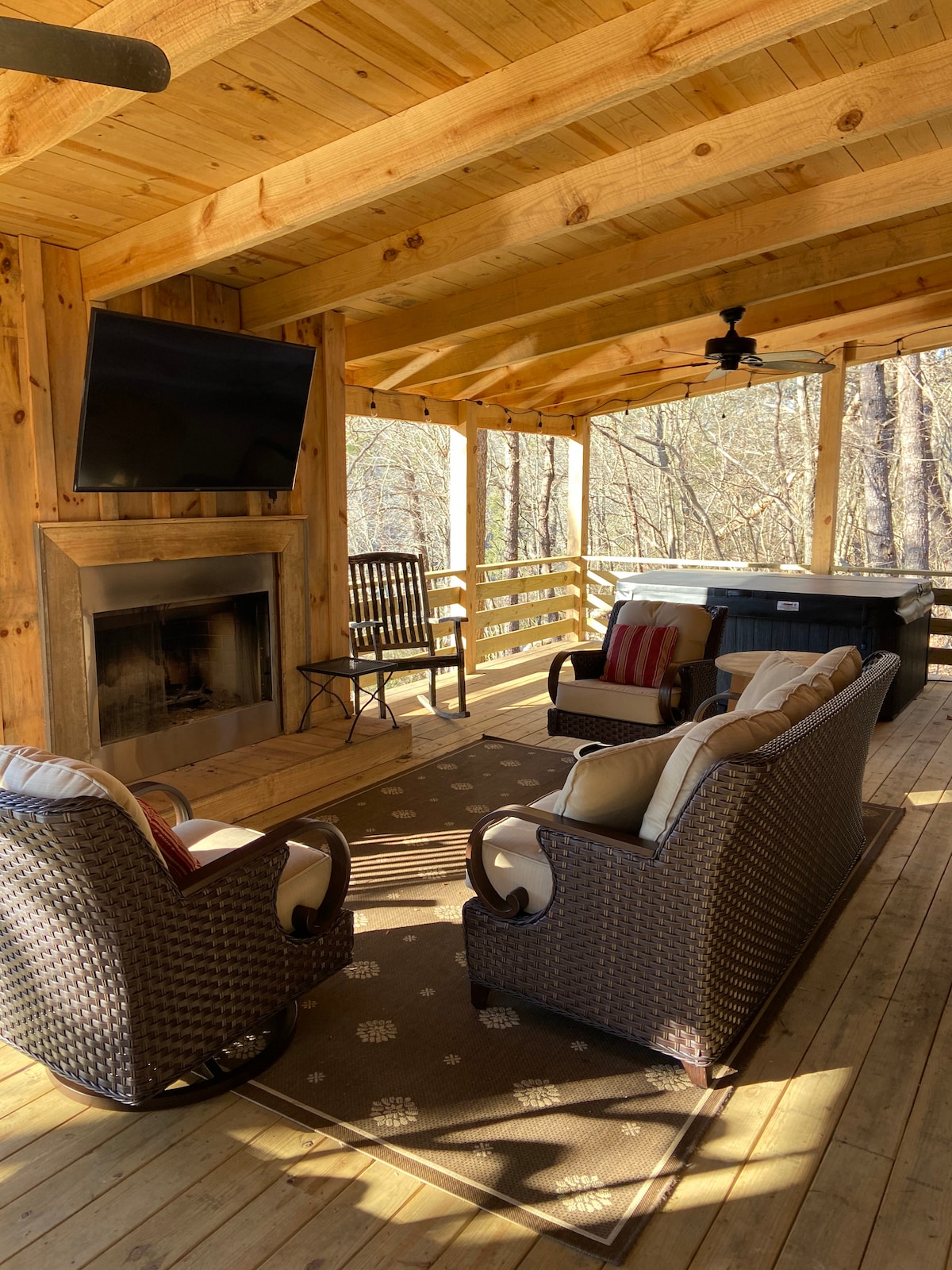 A spacious deck features comfortable seating arrangements, including two armchairs and a cozy sofa. A wood-burning fireplace is centrally located, with a large TV mounted above. A hot tub is positioned nearby, and the surrounding area is embraced by wooded views.
