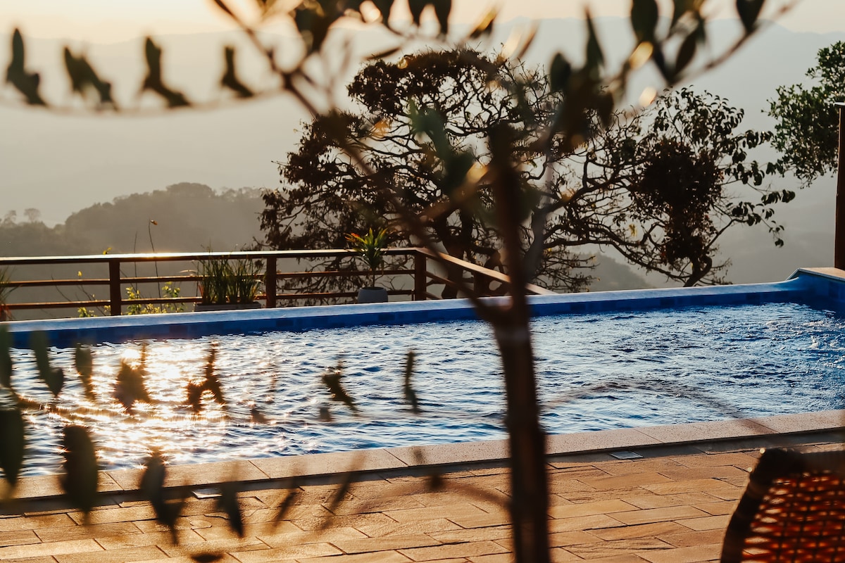 An infinity pool is set against a backdrop of softly lit hills during sunset, with reflections shimmering on the water's surface. Lush greenery frames the scene, enhancing the sense of tranquility in the outdoor space.