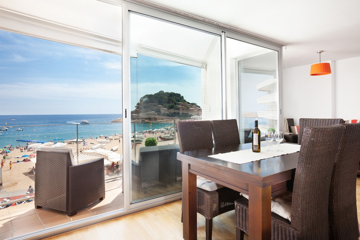 A dining area is visible, featuring a dark wooden table surrounded by six wicker chairs. Large glass doors open to reveal a scenic view of the beach and coastline, allowing natural light to fill the space. The table is set with a bottle of wine and glasses.