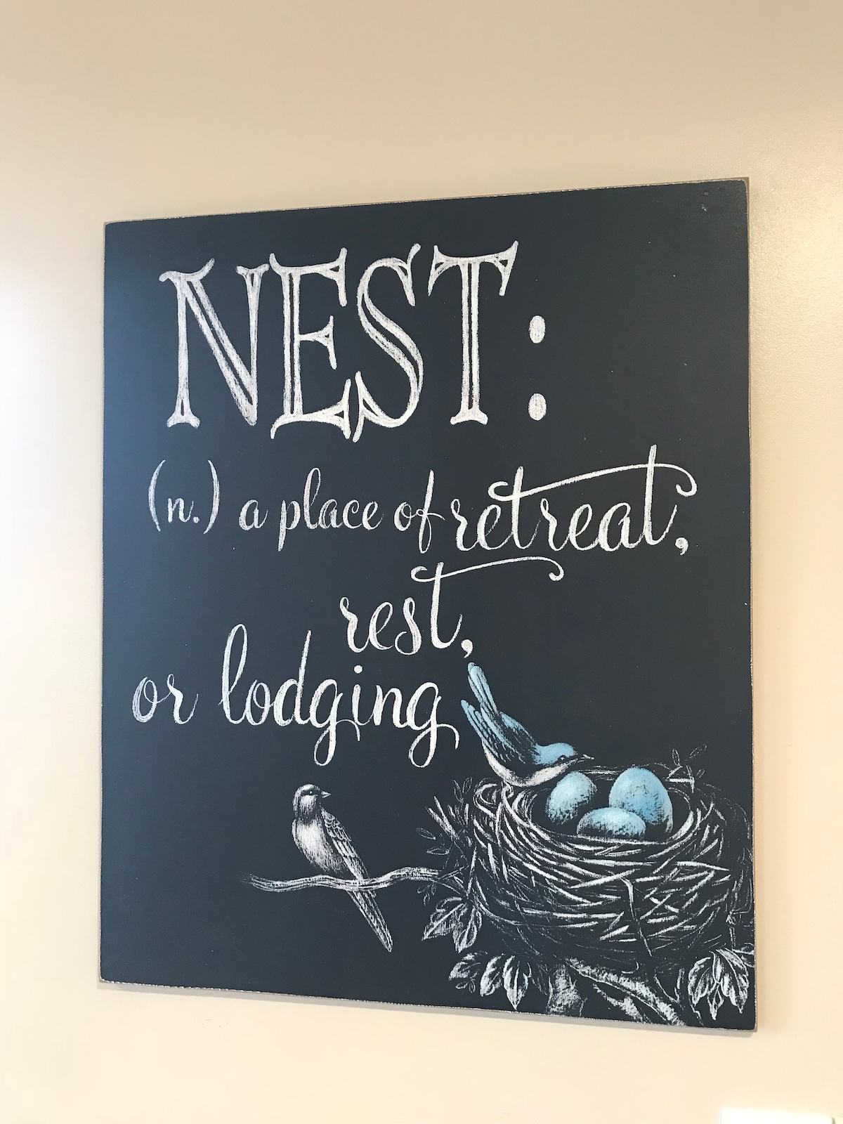 A decorative sign featuring the word 'NEST' is displayed on a wall. The definition describes it as 'a place of retreat, rest, or lodging,' alongside a drawing of a nest with blue eggs and a bird.