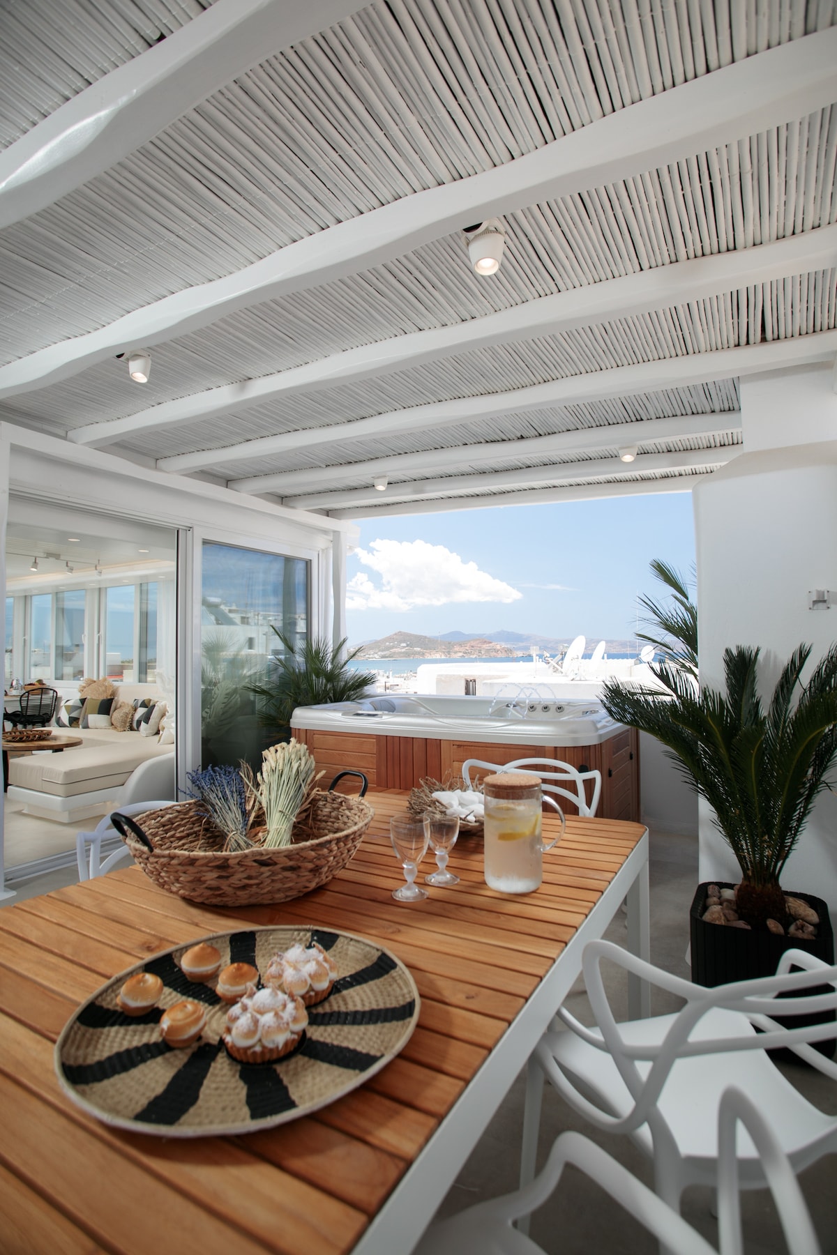An outdoor dining area features a wooden table adorned with a decorative woven basket and dessert treats. White chairs surround the table, complemented by greenery. Large glass doors open to the inviting space, offering views of the sea and bright blue sky beyond.