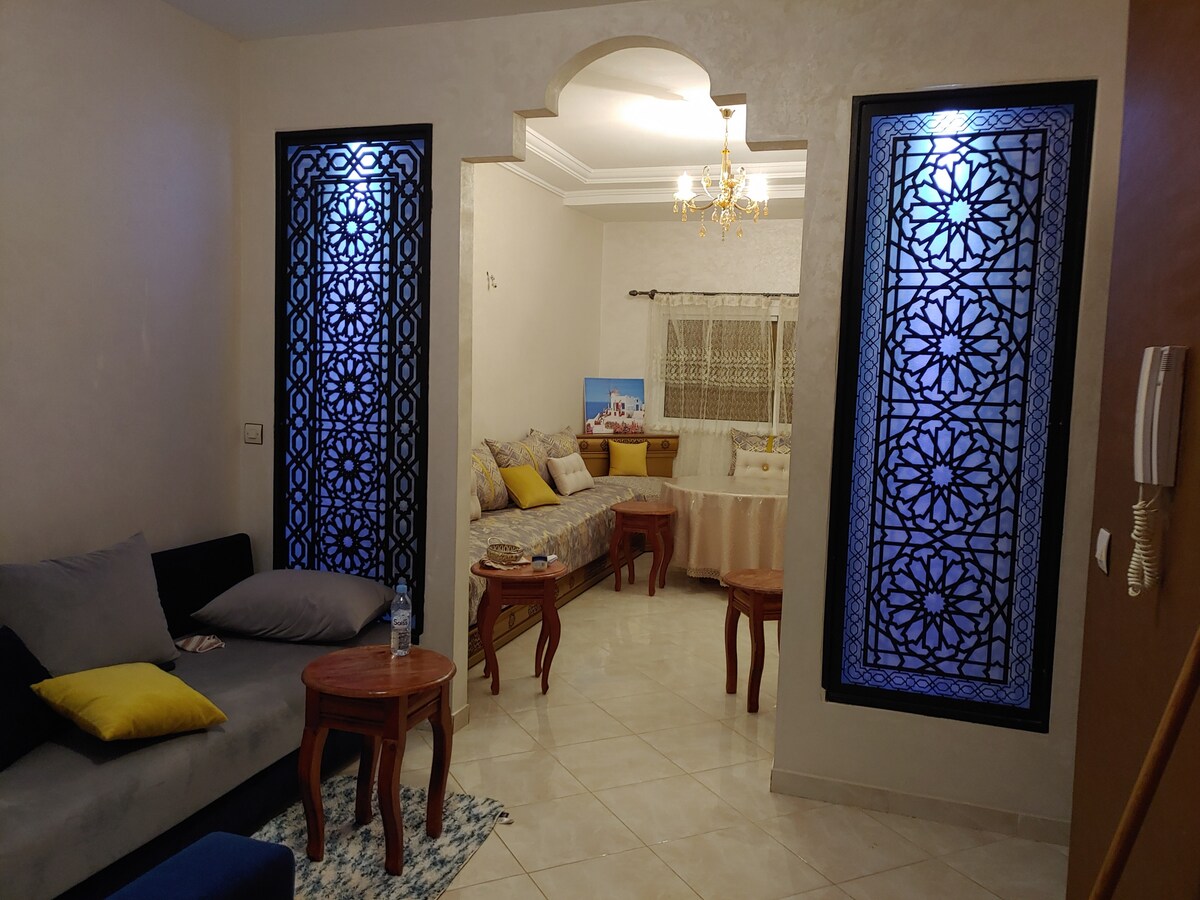 The living area features a cozy seating arrangement with dark cushions and decorative pillows, complemented by wooden tables. Intricate, patterned windows allow soft light to filter in, while a serene bedroom area is visible in the background, showcasing a comfortable bed and neutral decor.