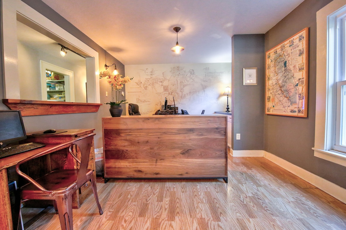 A welcoming front desk area features a wooden reception counter with a lamp and decorative flowers. A small desk with a laptop is positioned nearby. Light hardwood floors and neutral walls create an inviting atmosphere, complemented by natural light streaming through the window.