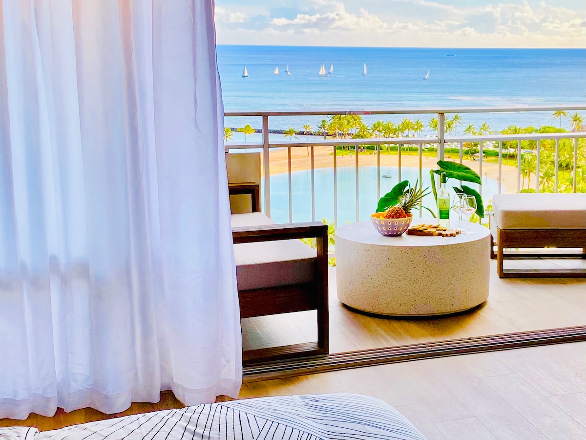 An expansive view of the ocean is visible through large white curtains. The outdoor balcony features a round stone table set with fresh fruit and light snacks, paired with two comfortable chairs. The tranquil sea and distant sailboats are framed by lush palm trees.