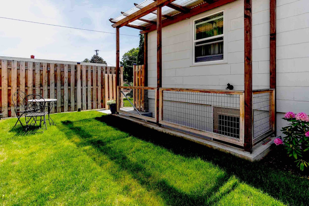 A grassy outdoor area is featured, accompanied by a small table and two chairs positioned under a wooden awning. The light-colored walls of the structure contrast with the green grass, while a fence provides privacy. Potted flowers add a gentle pop of color to the scene.