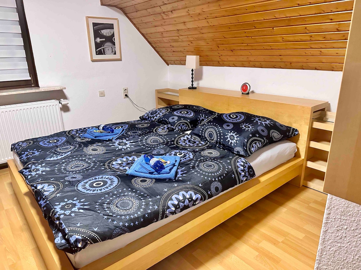 A spacious bed with a wooden frame is dressed in patterned bedding featuring circular designs. Two towels are neatly arranged at the foot of the bed. A small bedside table holds a lamp, while a picture hangs on the wall, enhancing the wood-paneled ceiling.