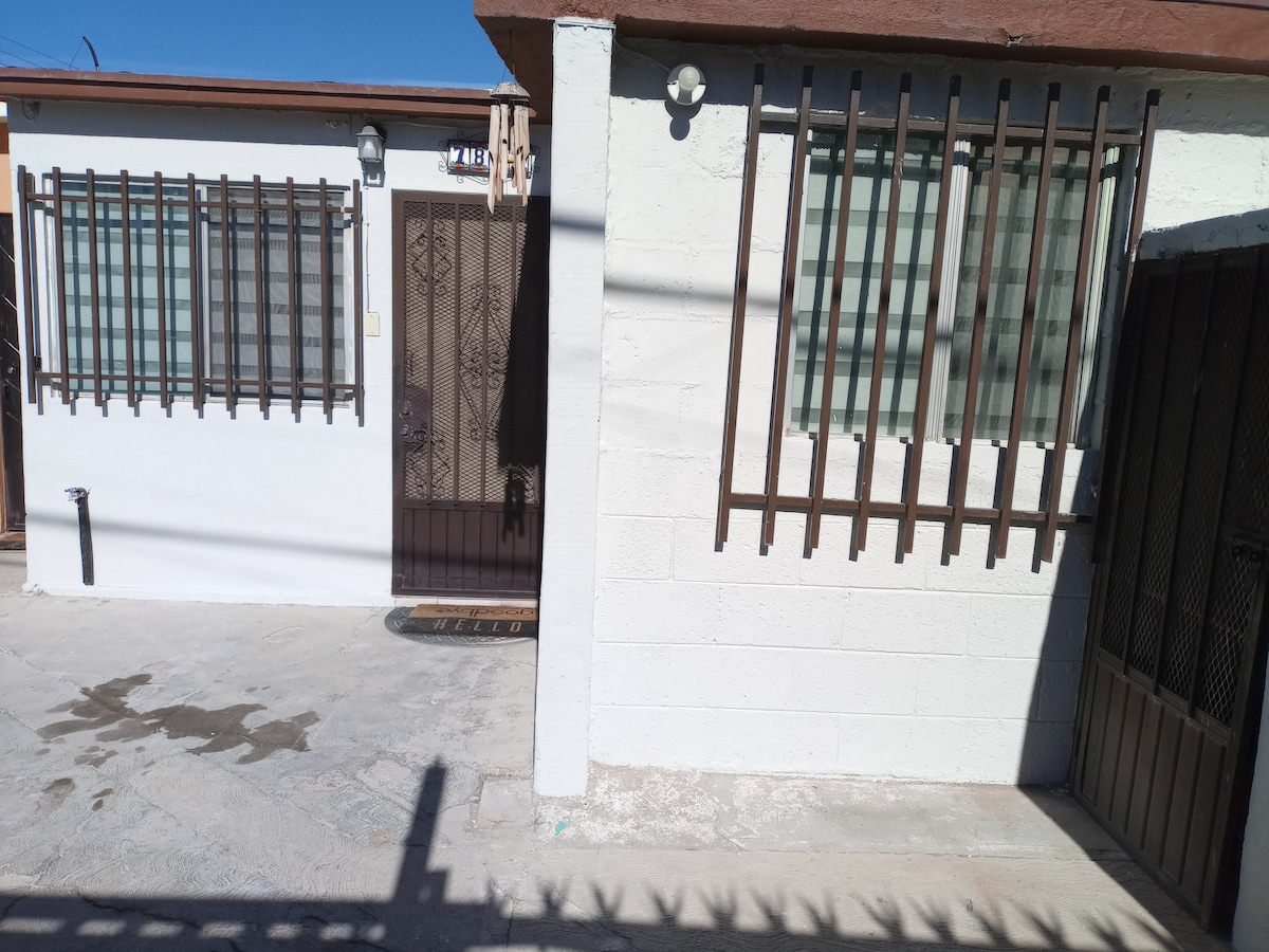 The exterior of the building features a simple, clean design with two windows adorned with protective bars. A solid door with a decorative grill provides entry. The area is well-lit and surrounded by a concrete pathway.