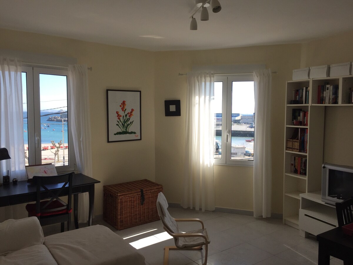 A sunlit room features two large windows offering views of the harbor. A wooden table with a chair is positioned next to the window, while a wicker storage box and an armchair provide additional seating. A bookshelf and a television are present on a white shelf.