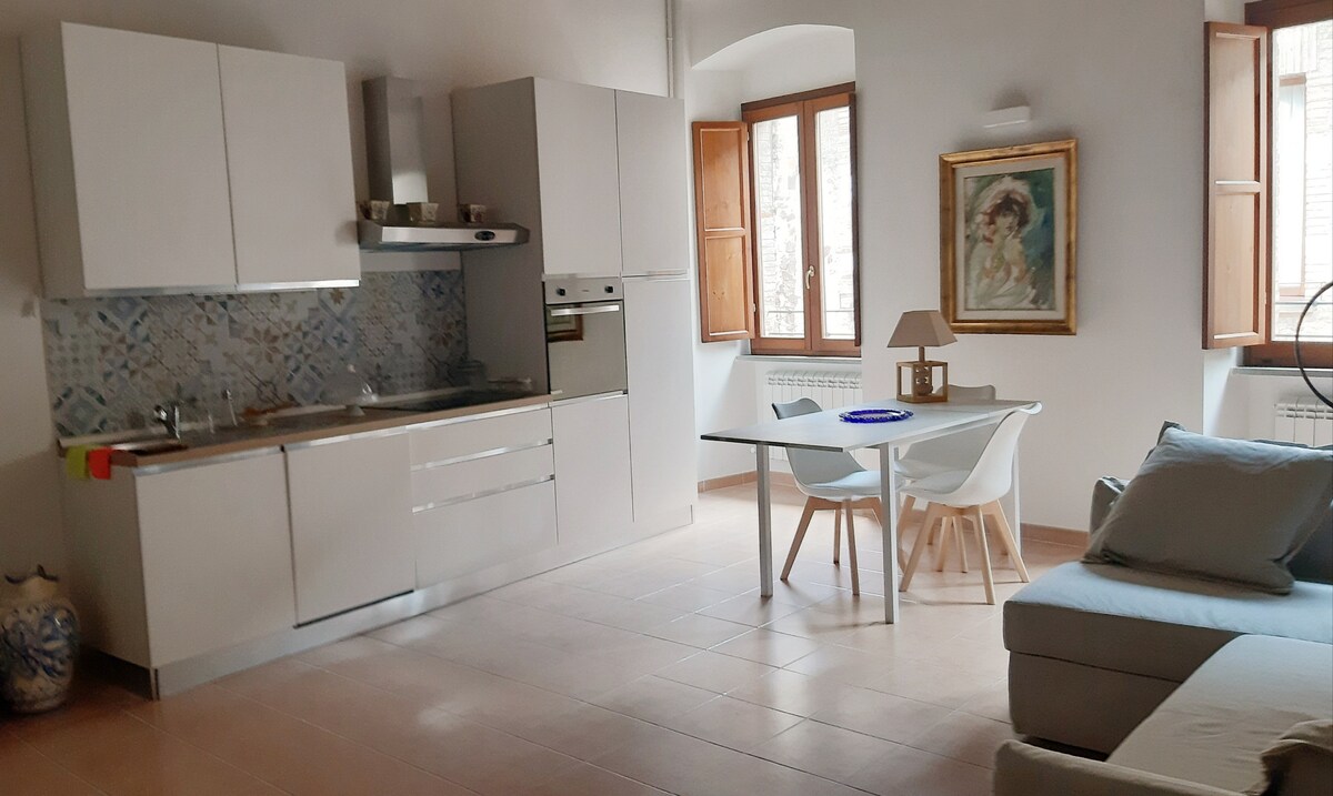 The kitchen area features modern cabinetry and a stylish backsplash. A dining table with four chairs and a lamp is positioned beside a cozy seating area. Natural light enters through windows with wooden shutters, enhancing the open layout of the space.